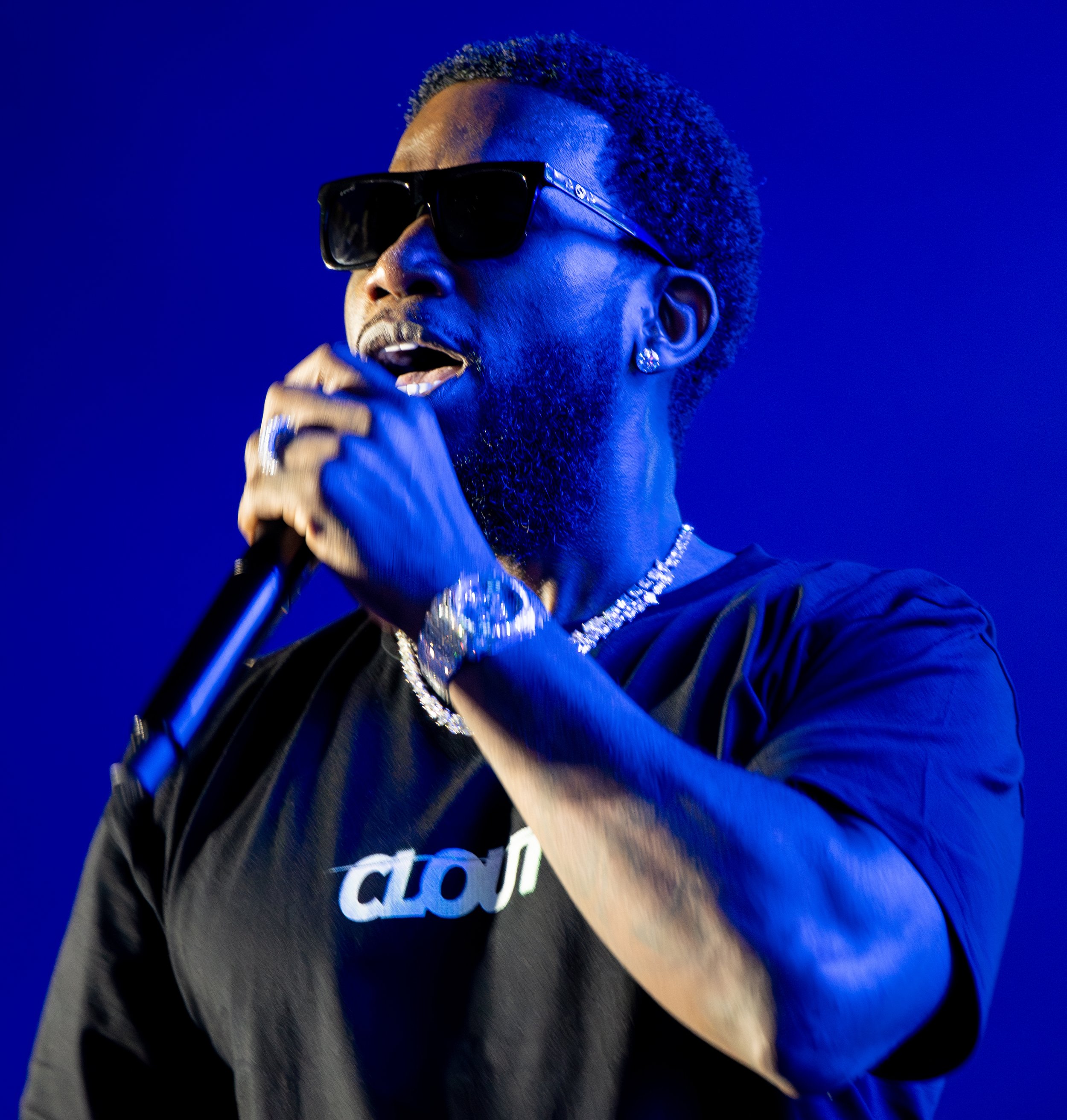 A person with short curly hair and sunglasses, singing into a microphone against a blue background, wearing a black shirt with white text and jewelry.