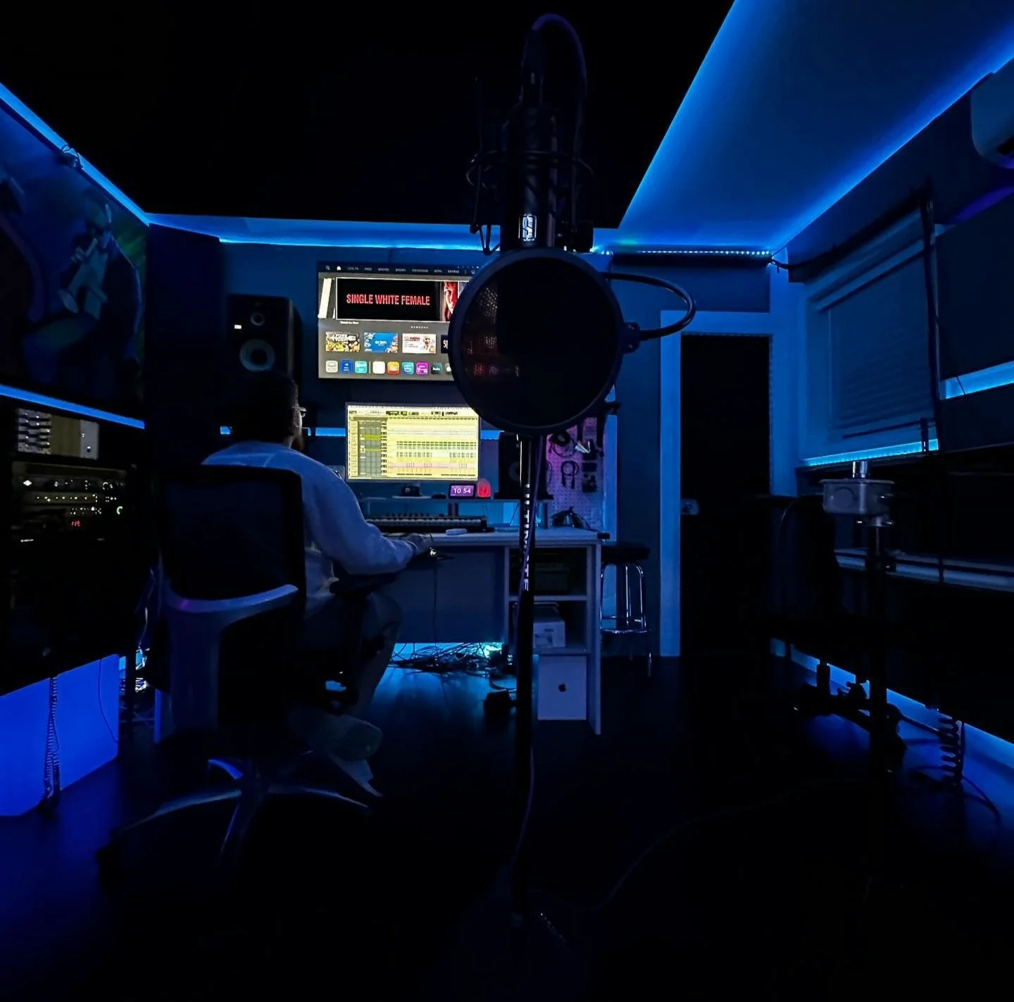 A recording studio with blue ambient lighting, a person sitting at a desk with multiple monitors and sound equipment, a microphone hanging from above, and a screen displaying various media options.