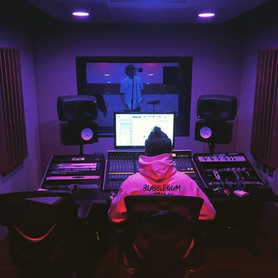 Music studio with person wearing a hoodie in front of mixing console, recording artist in booth, monitors, and sound equipment.