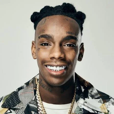Portrait of a smiling man with tattoos, wearing a patterned shirt and gold chains.