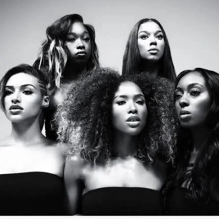 Six women with diverse hairstyles and expressions posing together in black tops, with a plain light background.