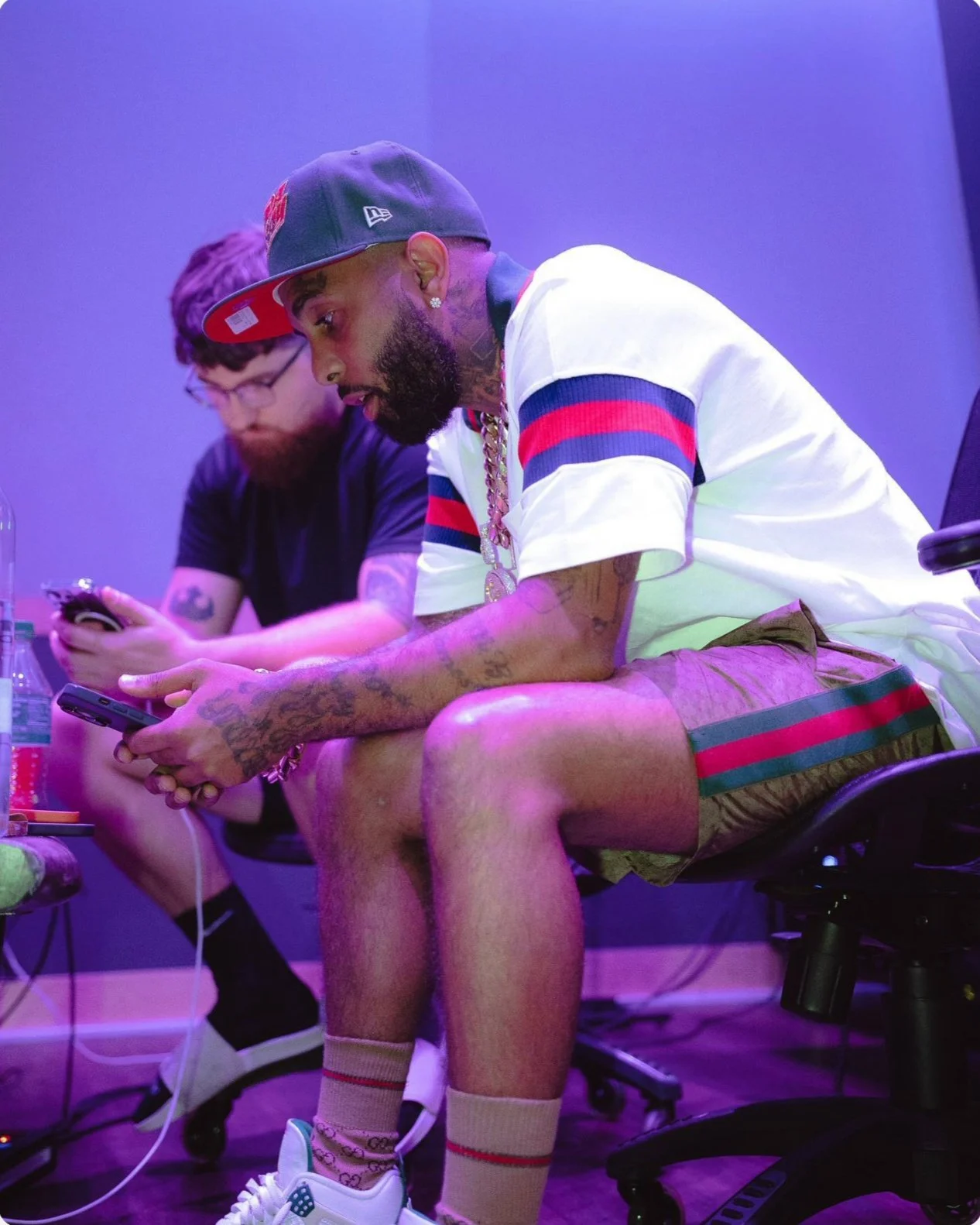 Two men sitting at a table, both looking at their phones, in a room with purple lighting. The man in front has tattoos, wears a cap, a white shirt with red and navy stripes, shorts, and sneakers, with a gold chain around his neck.