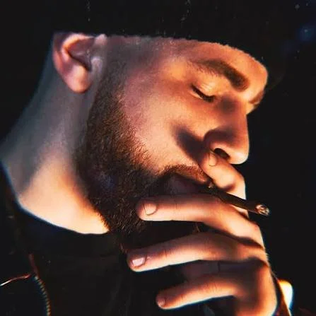 Close-up of a man with a beard and mustache, wearing a black beanie, smoking a cigarette or joint.