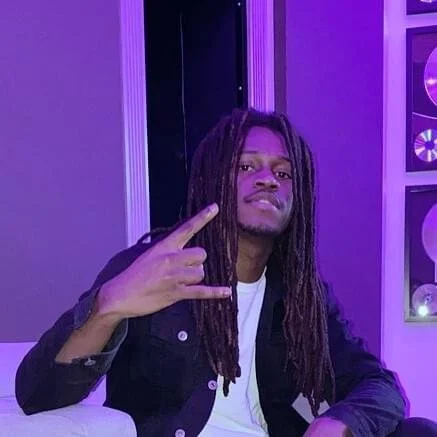 Man with long dreadlocks making a gesture with his hand indoors, with purple lighting.