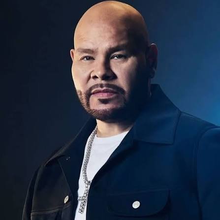 A bald man with a goatee and mustache wearing a dark jacket over a white shirt, posing against a dark background.