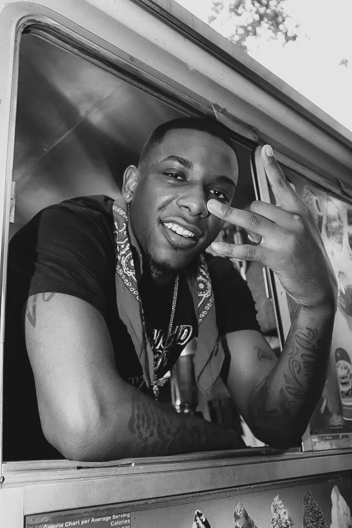 A young man with short hair and tattoos on his arms, smiling and making a gesture with his hand, leaning out of a food truck window.
