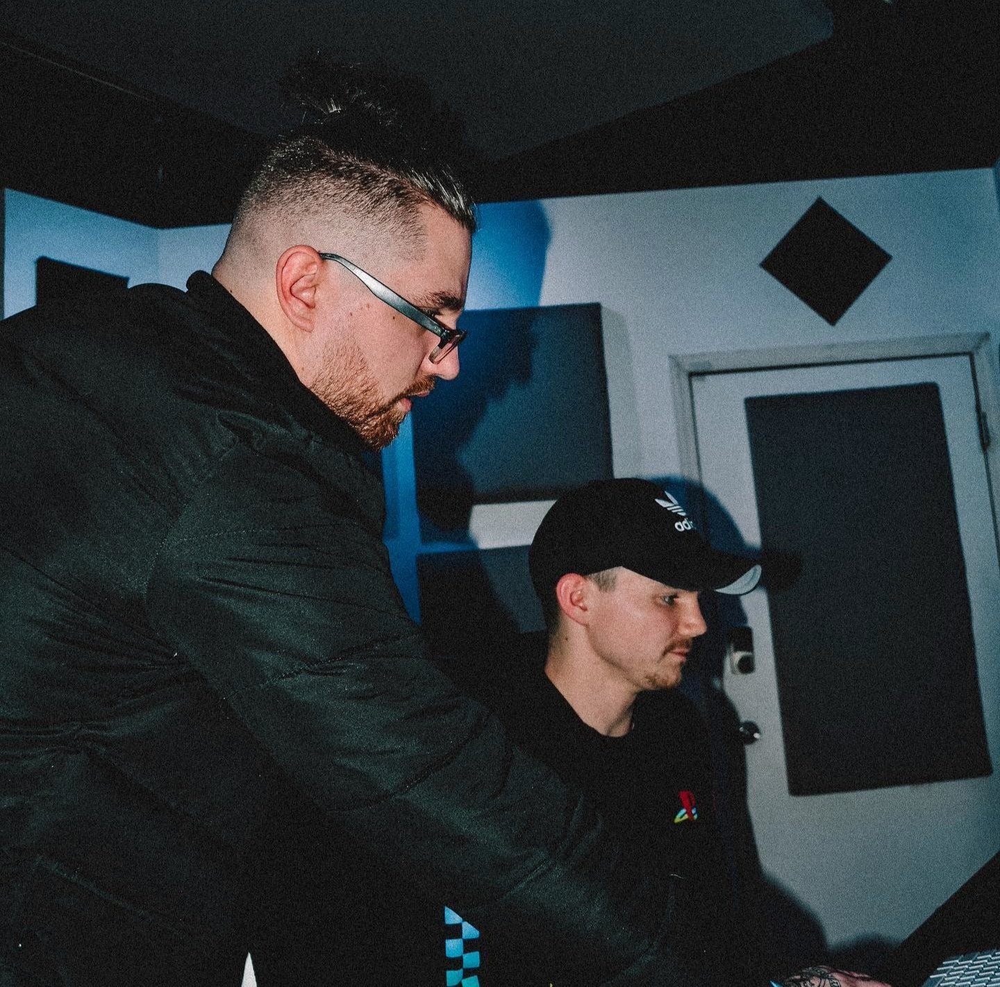 Two men working together in a music studio, one with glasses and a beard wearing a black jacket, and the other in a black cap with a PlayStation logo, focused on a sound mixing console.