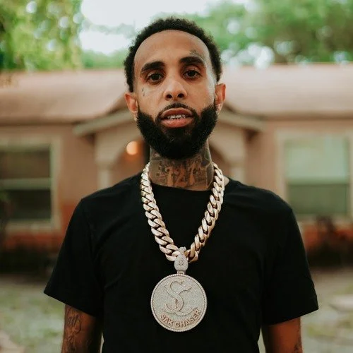 A man with a beard and tattoos wearing a black t-shirt and a large silver chain with a pendant, standing outdoors in front of a house.