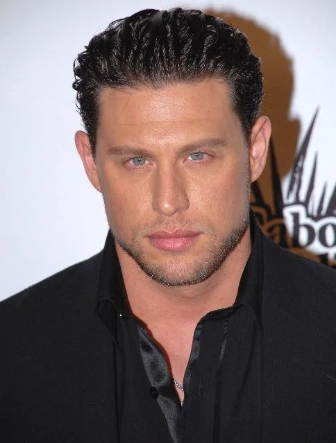 Close-up photo of a man with styled dark hair, blue eyes, and light stubble, wearing a black suit and shirt, standing in front of a blurred background with the American Idol logo.