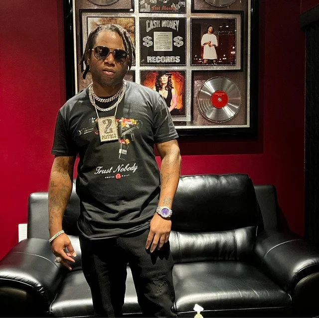 A man wearing sunglasses, a black t-shirt with slogan, jewelry, and a watch, standing in a music-themed room with framed records and photos on the wall, and a black leather couch.