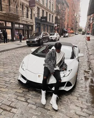 A person with curly hair sitting on the front of a white Lamborghini parked on a cobblestone city street, with buildings and pedestrians visible in the background.