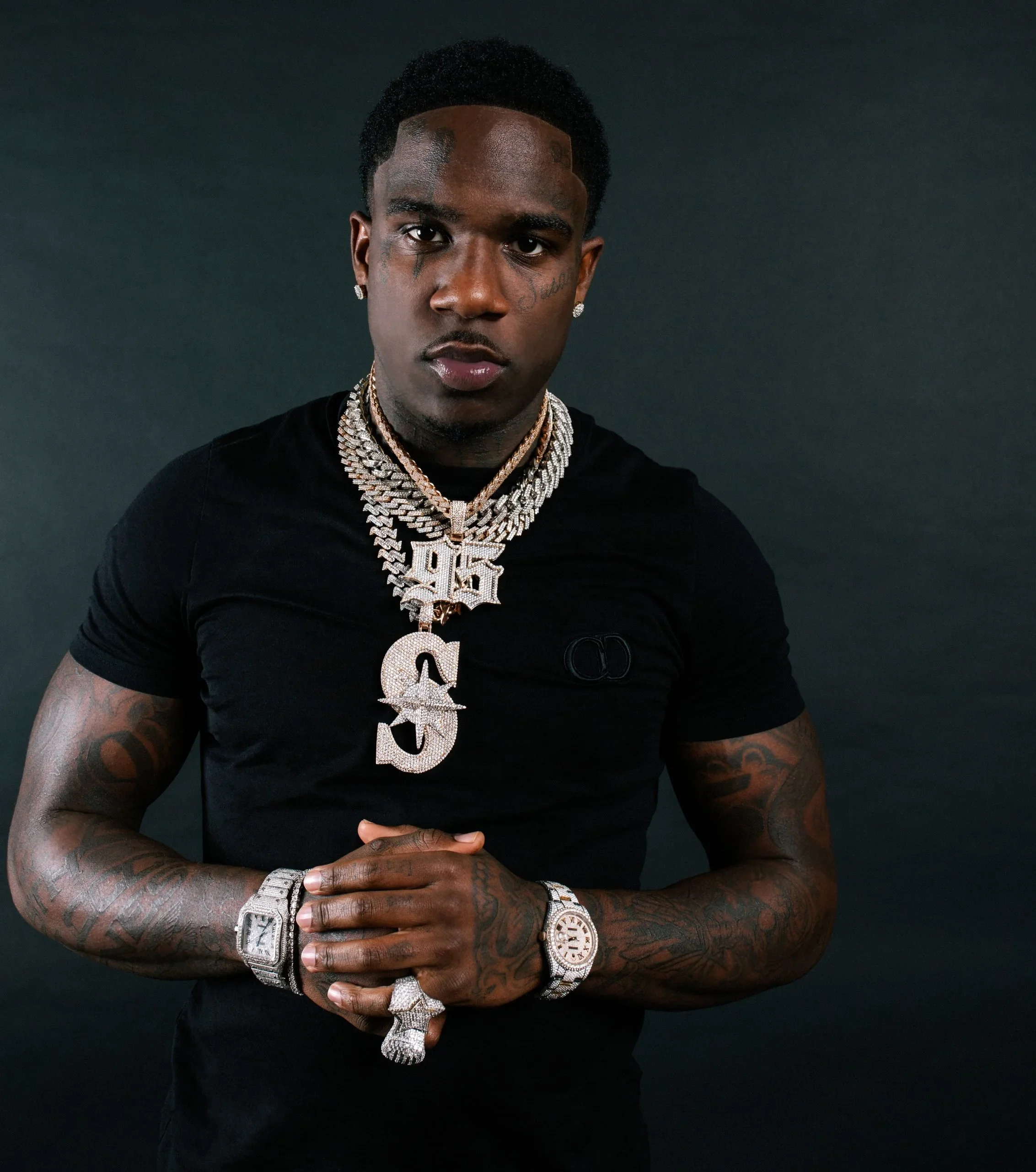 A man with tattoos, wearing multiple gold chains and diamond jewelry, including a watch, bracelet, and ring, stands against a dark background.