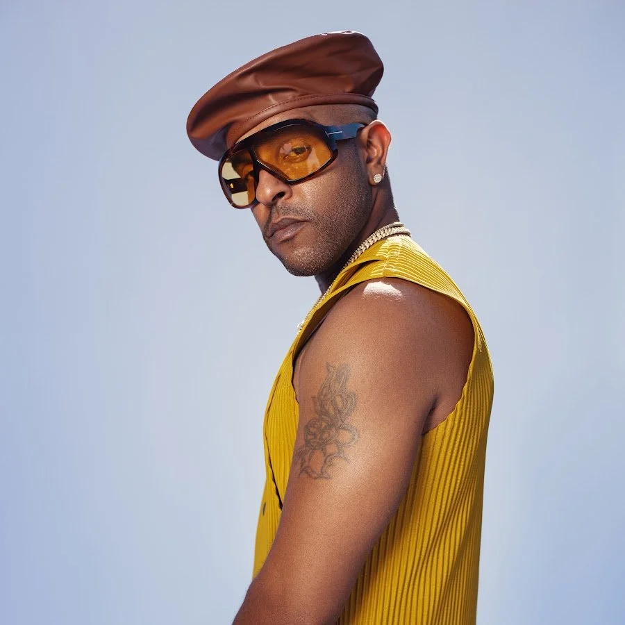 Man with tan skin wearing large glasses, a brown leather cap tilted to the side, a yellow sleeveless shirt, and a silver chain necklace, posing against a light blue background.