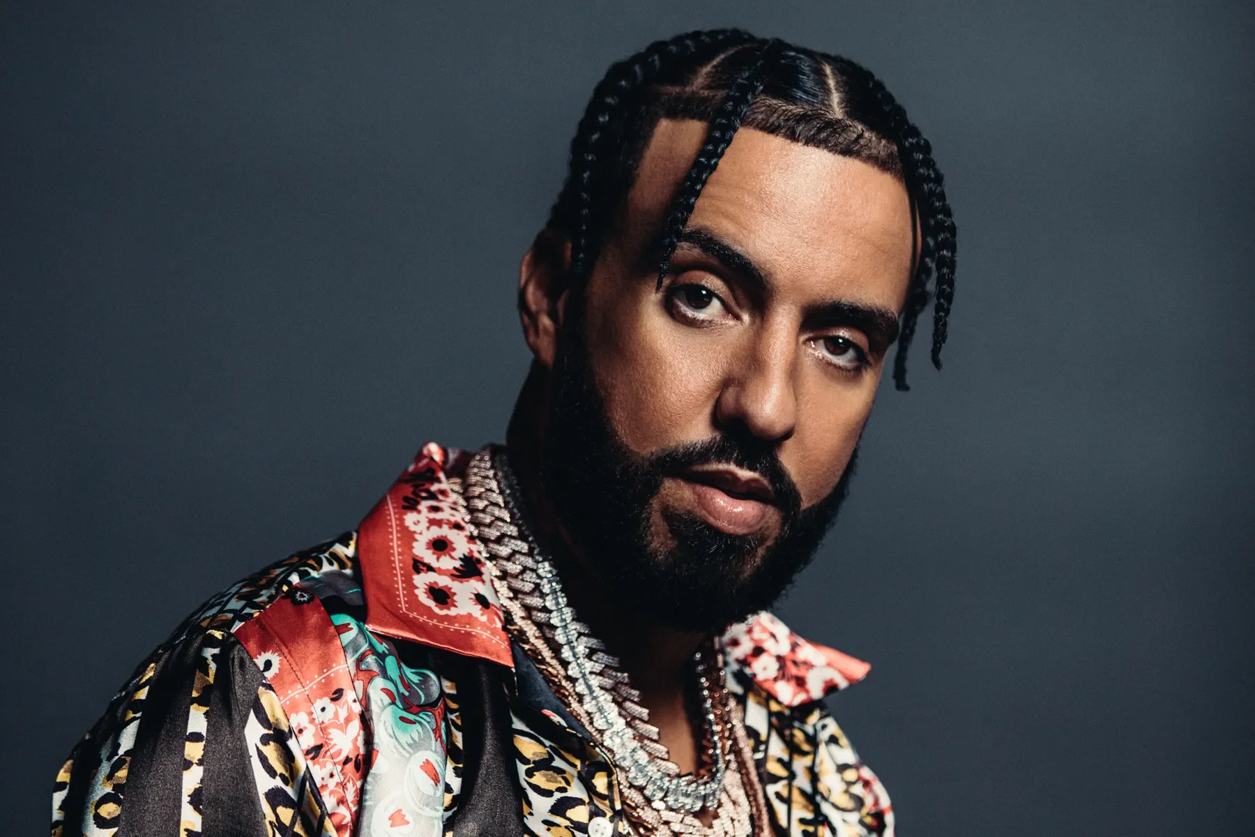 Close-up portrait of a man with braided hair, a beard, wearing colorful patterned clothing and multiple silver necklaces against a dark background.
