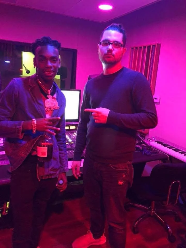 Two men in a recording studio with music equipment and colorful lighting. The man on the left is holding a drink and making a peace sign, while the man on the right is pointing at him.