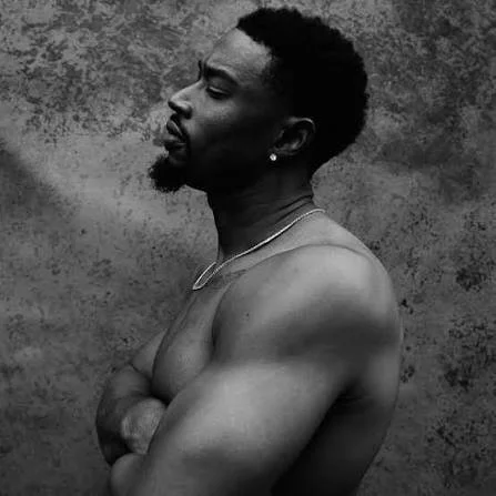 A shirtless man with a muscular build and styled hair, standing against a textured background, looking to the side.
