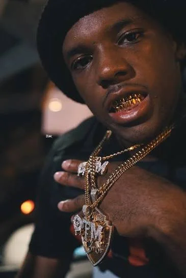 Young man wearing gold teeth, gold chains, and a black hat, looking at the camera with a serious expression, with his hand on his chest in a close-up shot.