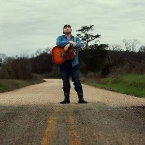 A man standing in the middle of a rural dirt road, playing an acoustic guitar, with trees and cloudy sky in the background.
