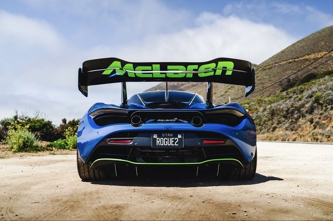 Blue McLaren sports car with a large rear wing and custom license plate reading ROGUE2, parked on a dirt road with hills and clear skies in the background. RG Motors McLaren
