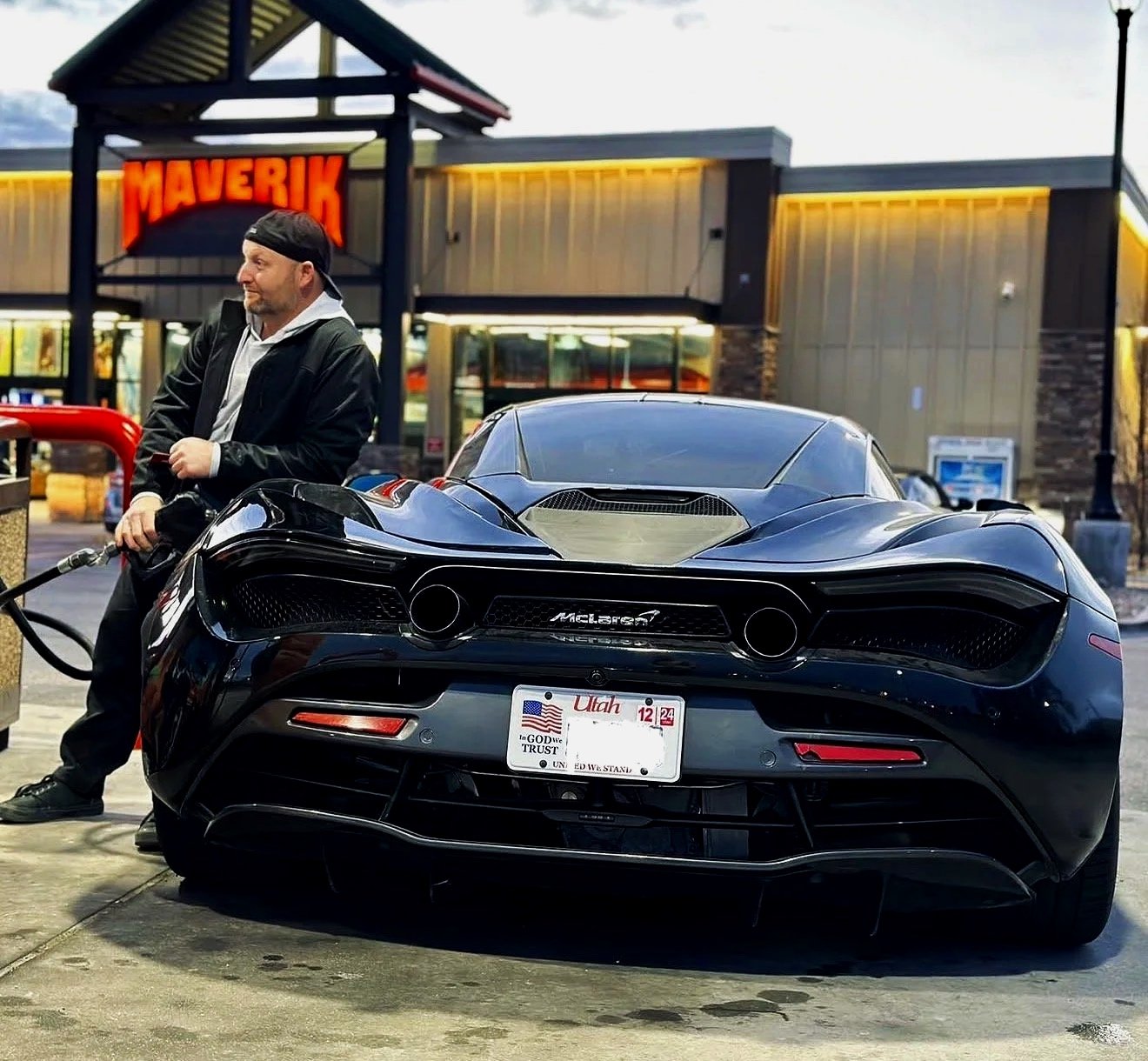 A man in a black jacket and cap fueling a black RG Motors McLaren sports car at a gas station in front of a store with a large red "Maverik" sign in Utah