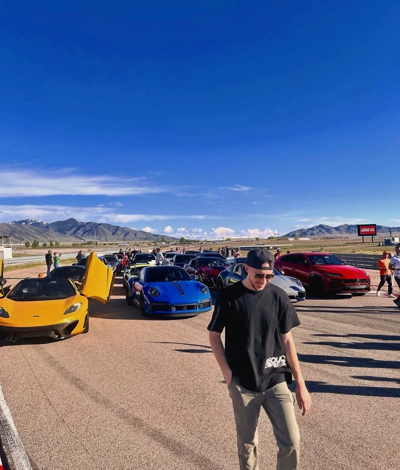 A race track filled with colorful sports cars, with an Ethan Ashby wearing sunglasses and a black baseball cap walking in front. In the background, there are mountains and a blue sky, with several people standing around and a large red billboard. A