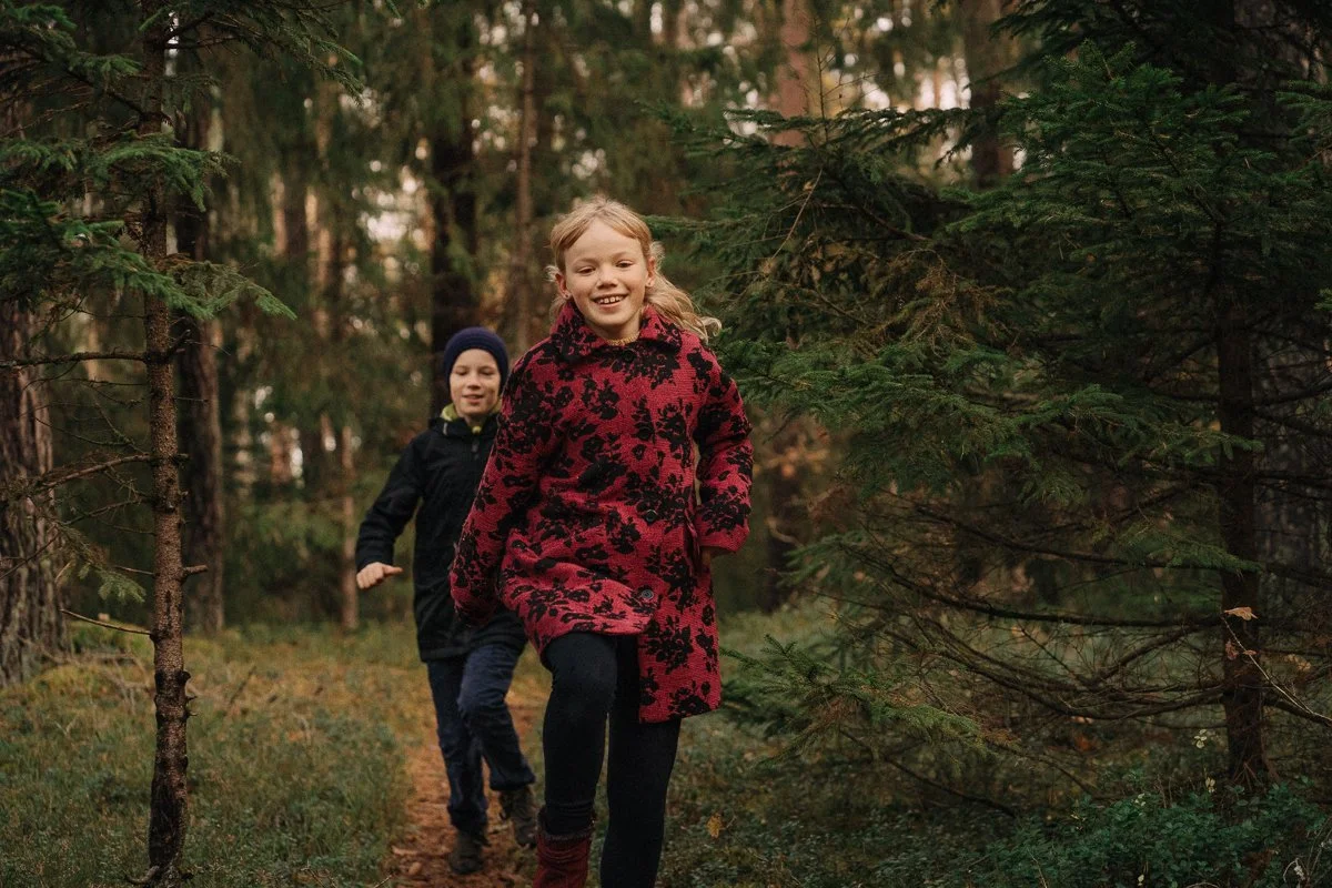 Two children, a girl in a red and black patterned coat and a boy in a black jacket and hat, are running along a forest trail among pine trees.