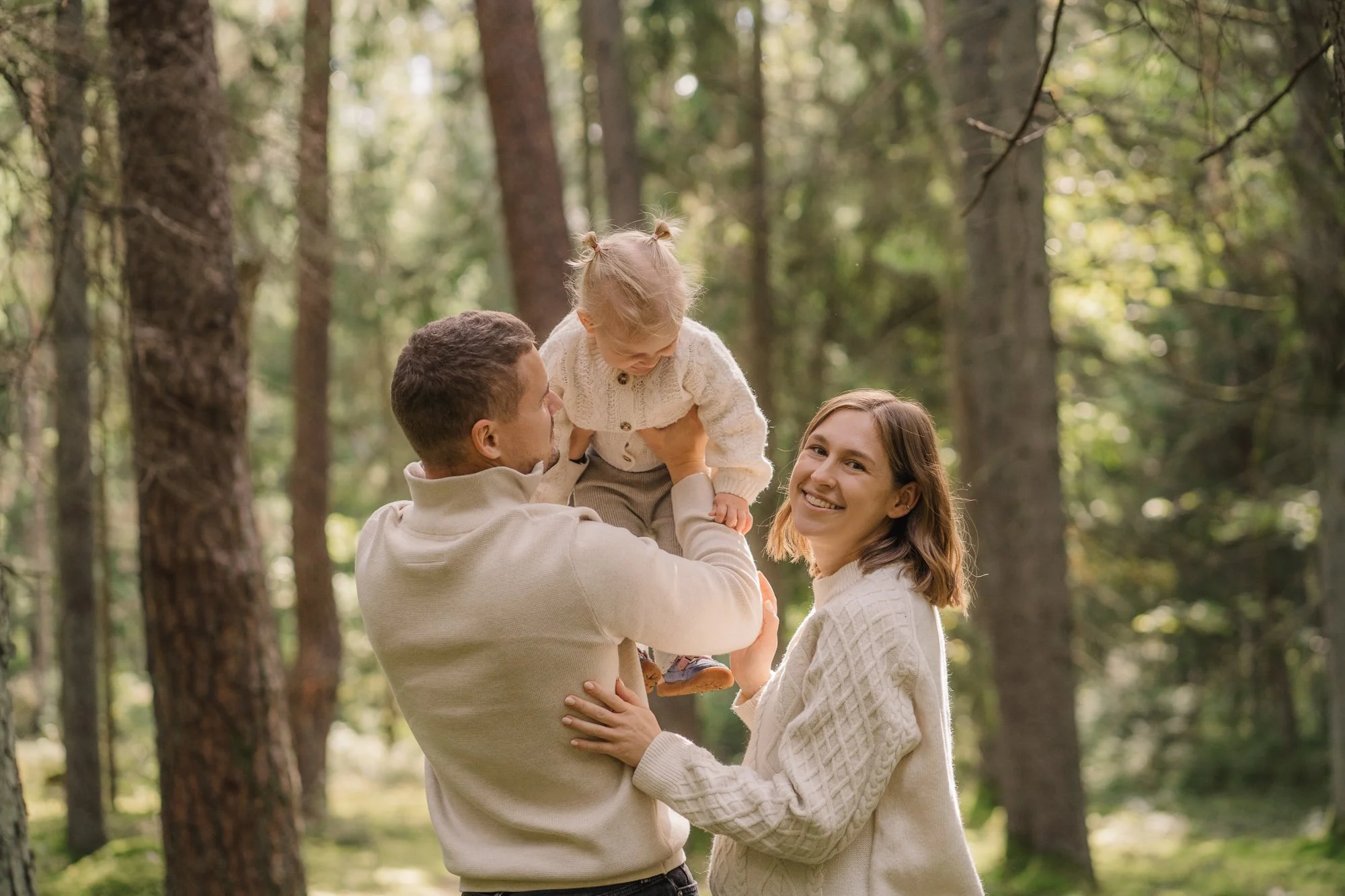 A man holding a young girl up in the air while a woman stands nearby smiling in a forested area.