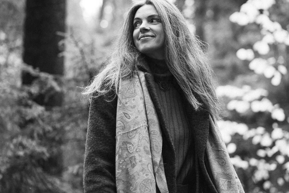 A woman with long, wavy hair is outdoors in a forest, wearing a coat and scarf, looking to the side with a smile.