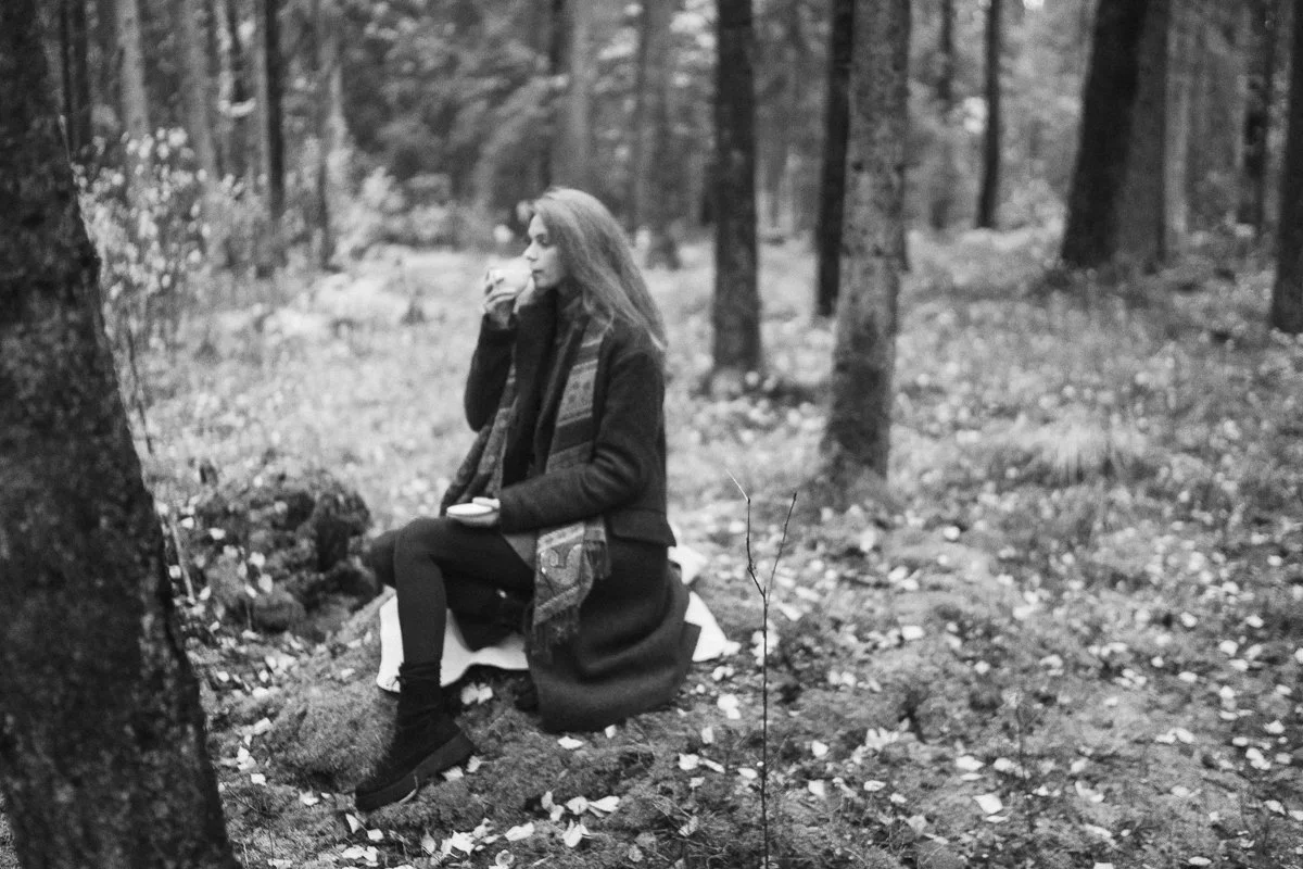 A woman sitting in a forest, wearing a jacket and scarf, holding a cup, with trees and fallen leaves around her in black and white.