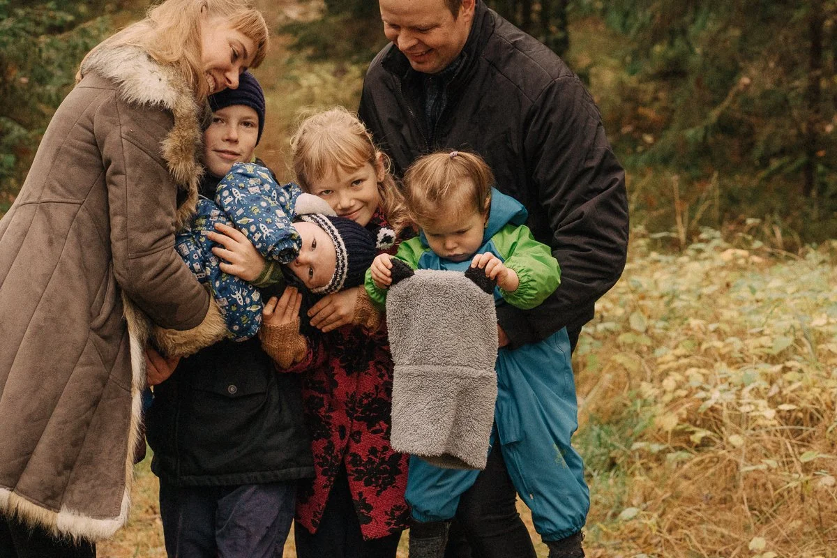 A family of five, including two adults and three children, are outdoors in a wooded area, dressed warmly for fall, gathered close together with smiles and affection.