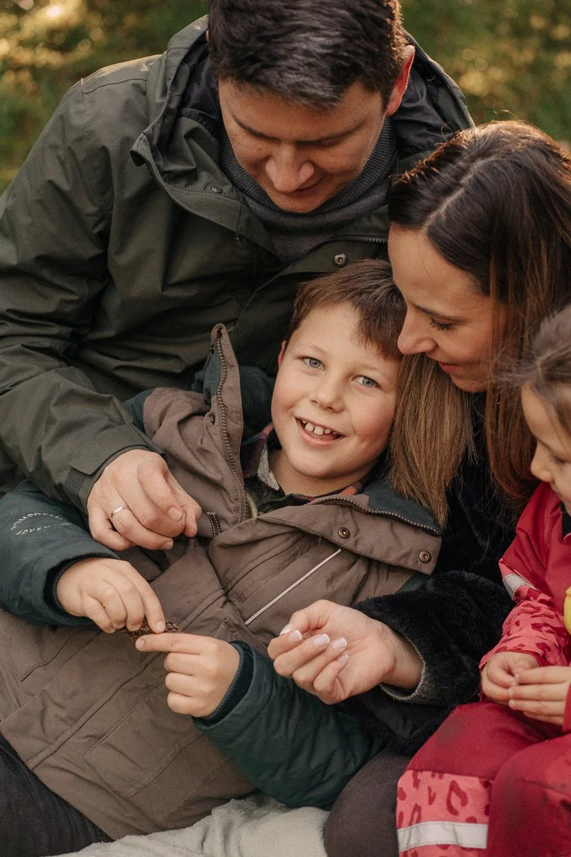 Family of four, including a smiling young boy, enjoying a moment outdoors during fall, surrounded by trees with orange leaves.
