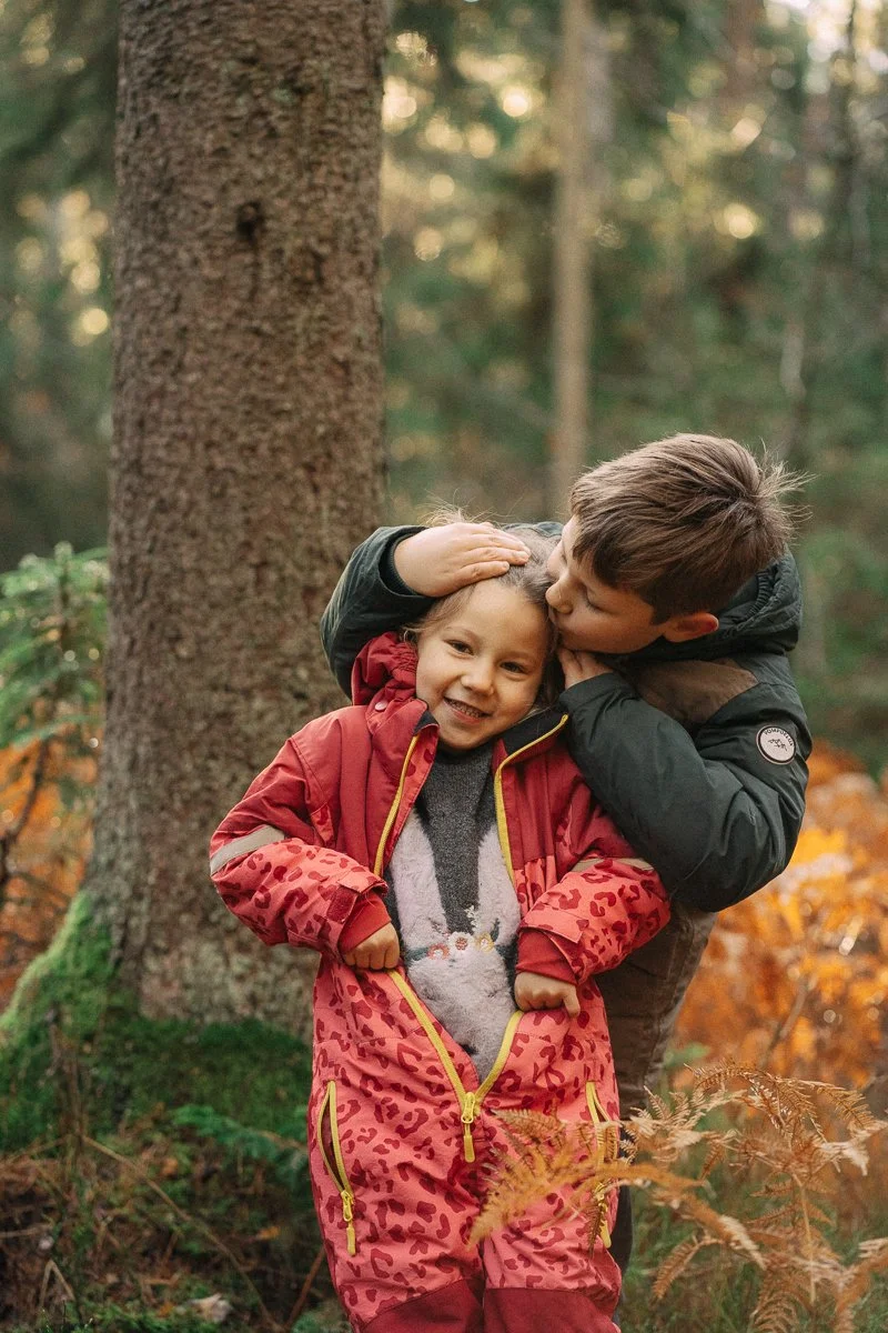 A young boy kisses a girl on the cheek while hugging her in a forest with autumn leaves.