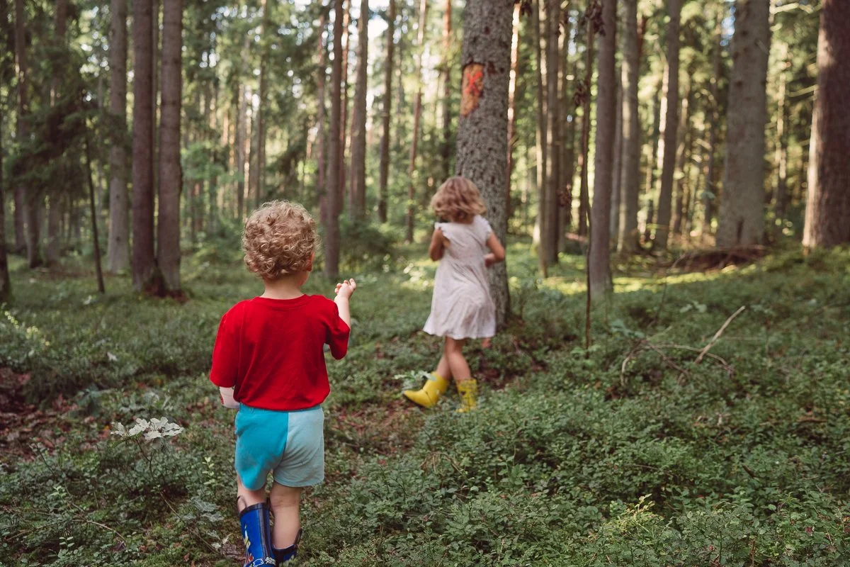 Two children, a boy and a girl, walking through a forest; the boy is wearing a red shirt, blue shorts, and rain boots, while the girl is wearing a white dress and yellow rain boots.