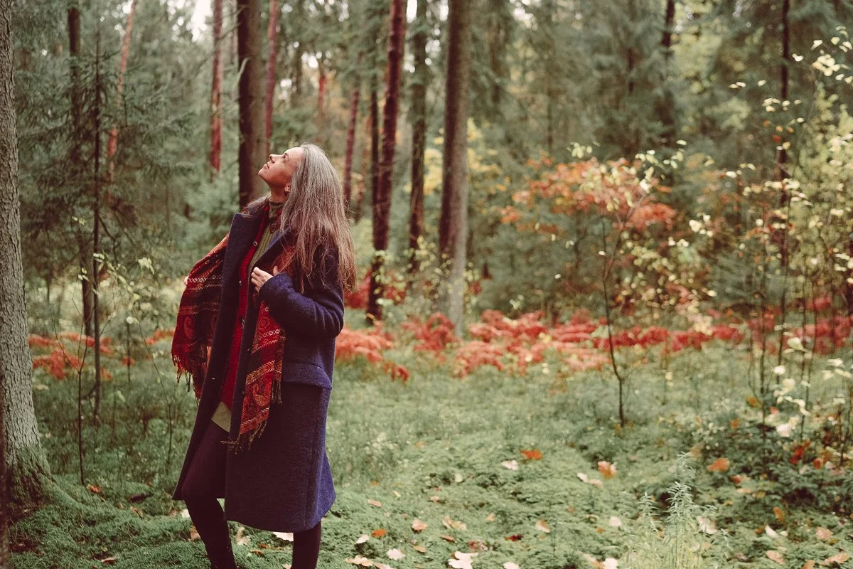 A woman with gray hair standing in a forest during fall, wearing a coat and scarf, looking up and smiling.