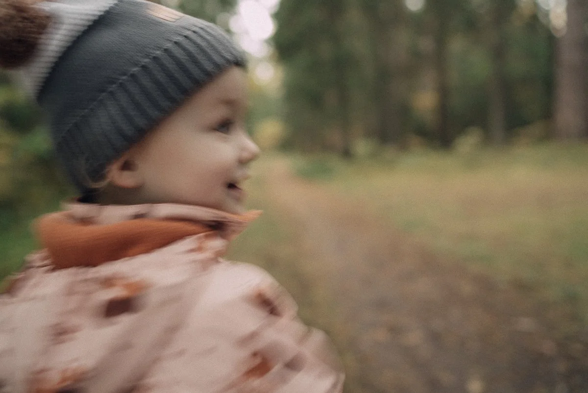A young child with a gray knit hat and pink jacket running outdoors in a wooded area.