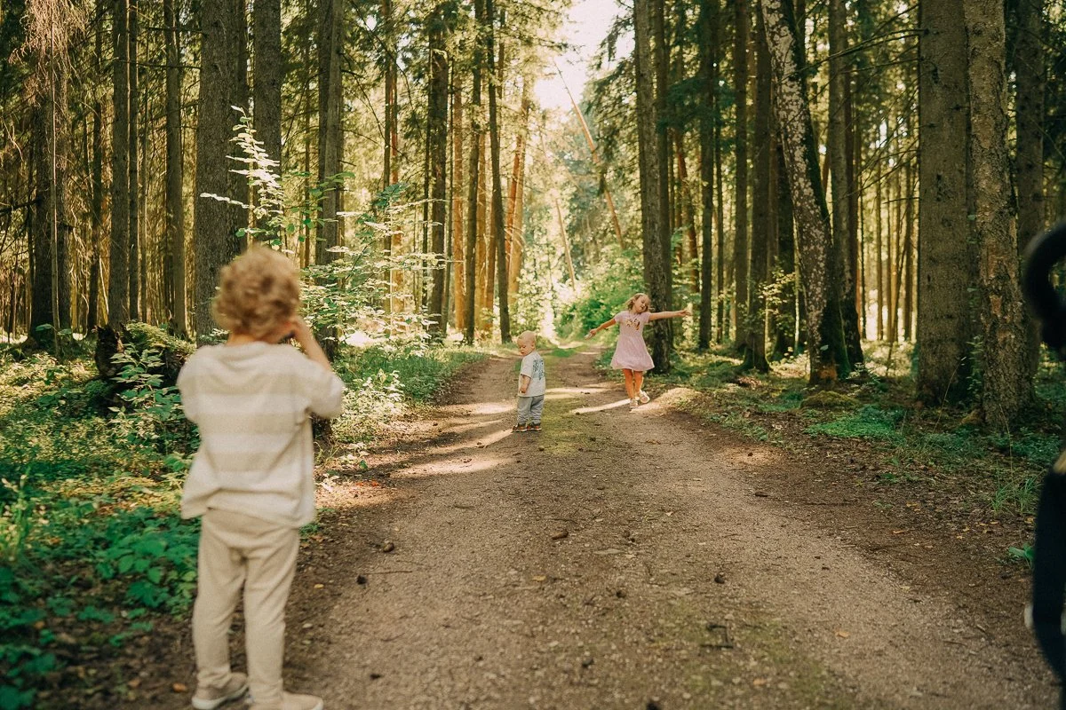 Children playing and posing for a photo on a forest trail with tall trees and sunlight filtering through the canopy.