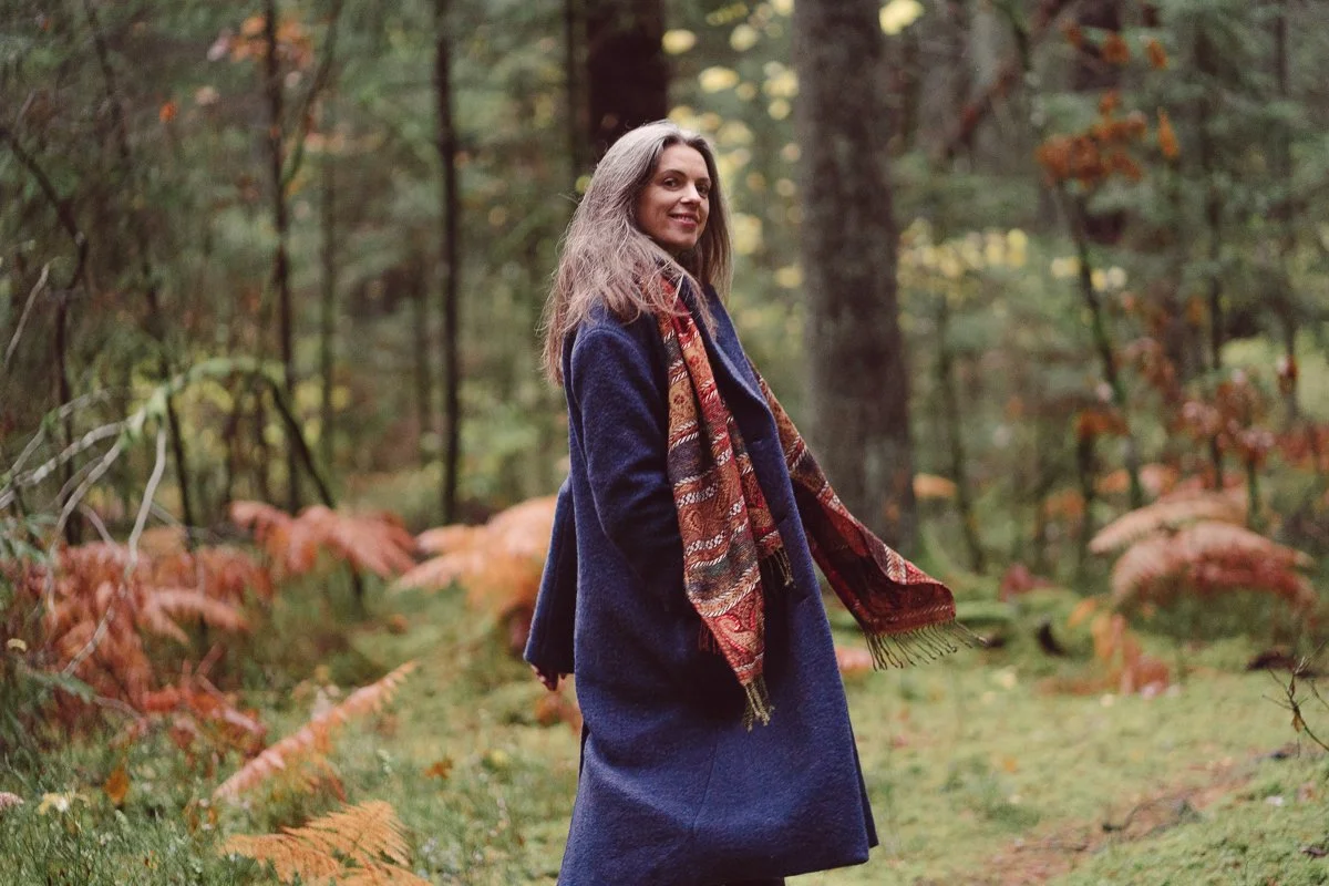 A woman in a blue coat and patterned scarf walking in a forest with autumn foliage.