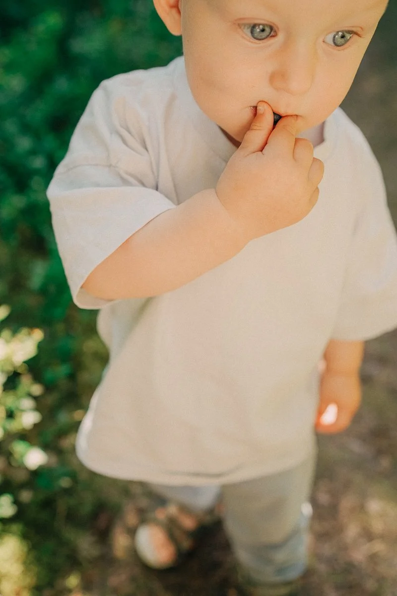A young child with blue eyes and fair skin, standing outdoors on a dirt path, with green foliage in the background, wearing a white shirt and gray pants, with one hand near their mouth.