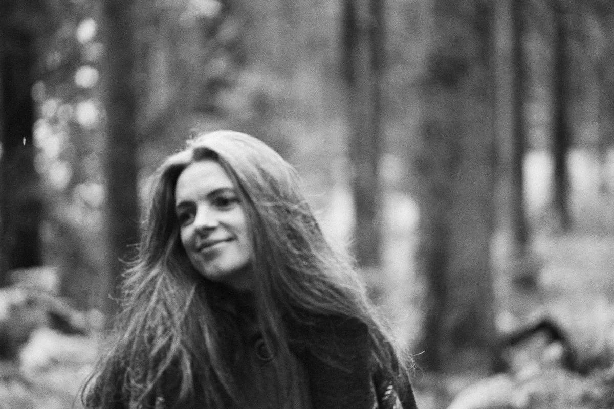 A black and white photo of a young woman with long hair smiling outdoors in a forest.