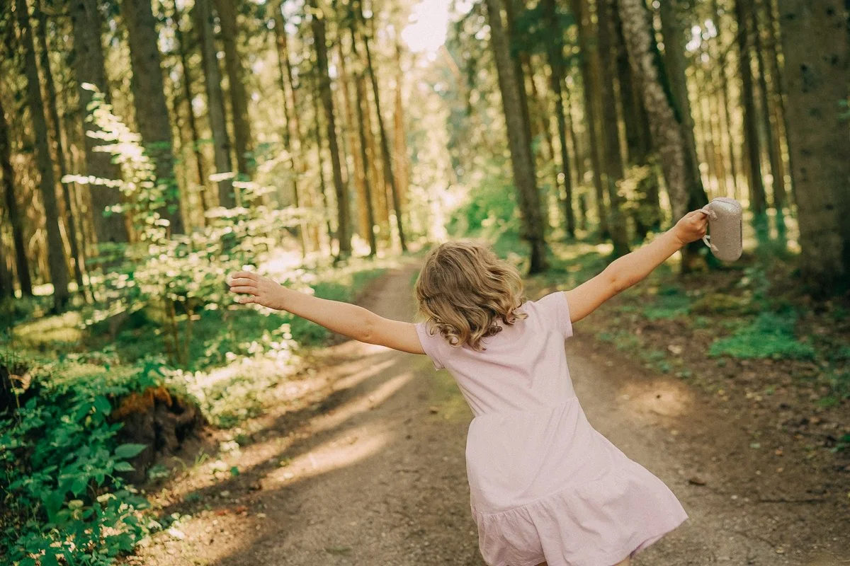 A young girl with curly hair wearing a pink dress is joyfully spinning on a dirt trail in a sunlit forest, holding a gray water bottle in her right hand with arms outstretched.