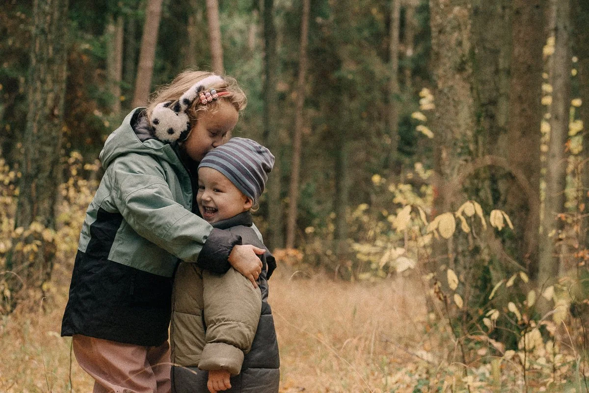 A girl with earmuffs hugging a young boy in a forest with yellow and green leaves
