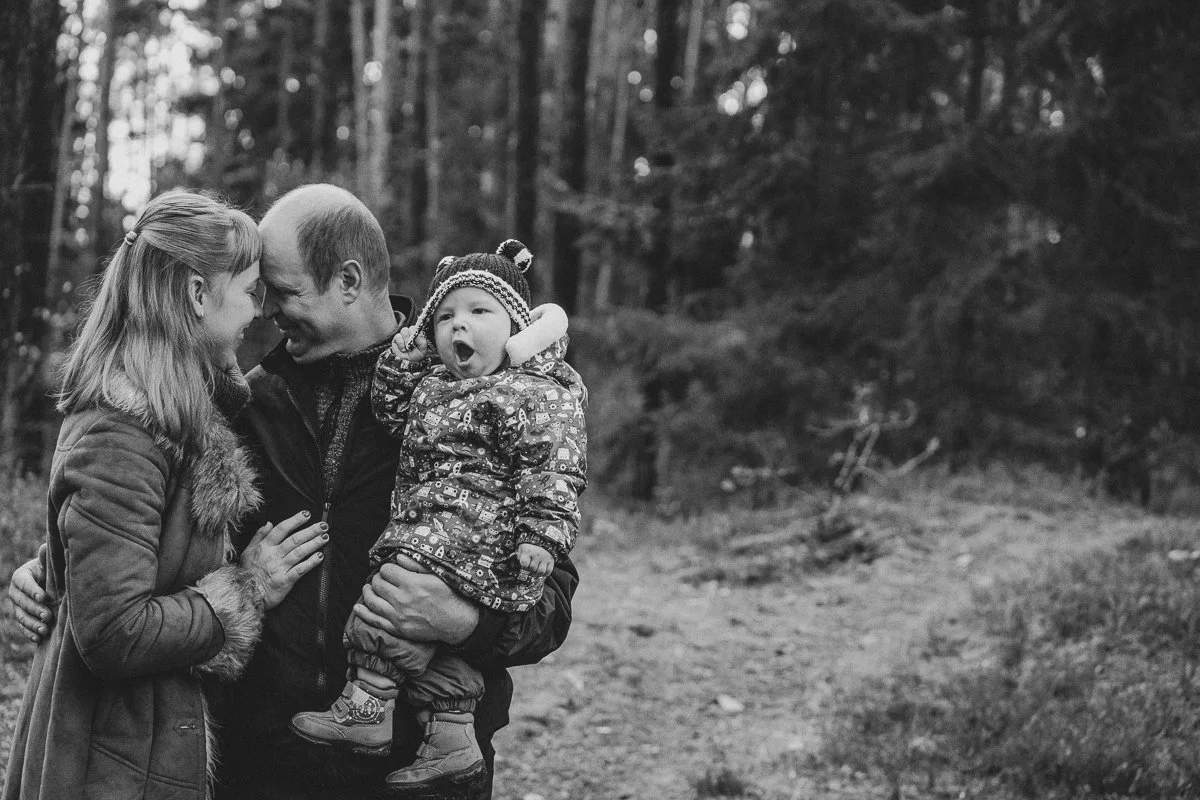 Black and white photo of a family of three outdoors in a forest. The father is holding a young girl, and they are all touching foreheads, smiling. The girl is wearing a winter hat and patterned jacket, and the mother is wearing a jacket with fur trim