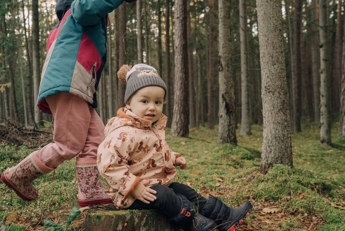 A young child sitting on a tree stump in a forest, wearing a beige jacket with a squirrel pattern, black pants, and boots, with a gray knit hat featuring a character, and an older child standing behind, partly out of the frame, wearing pink pants, fl