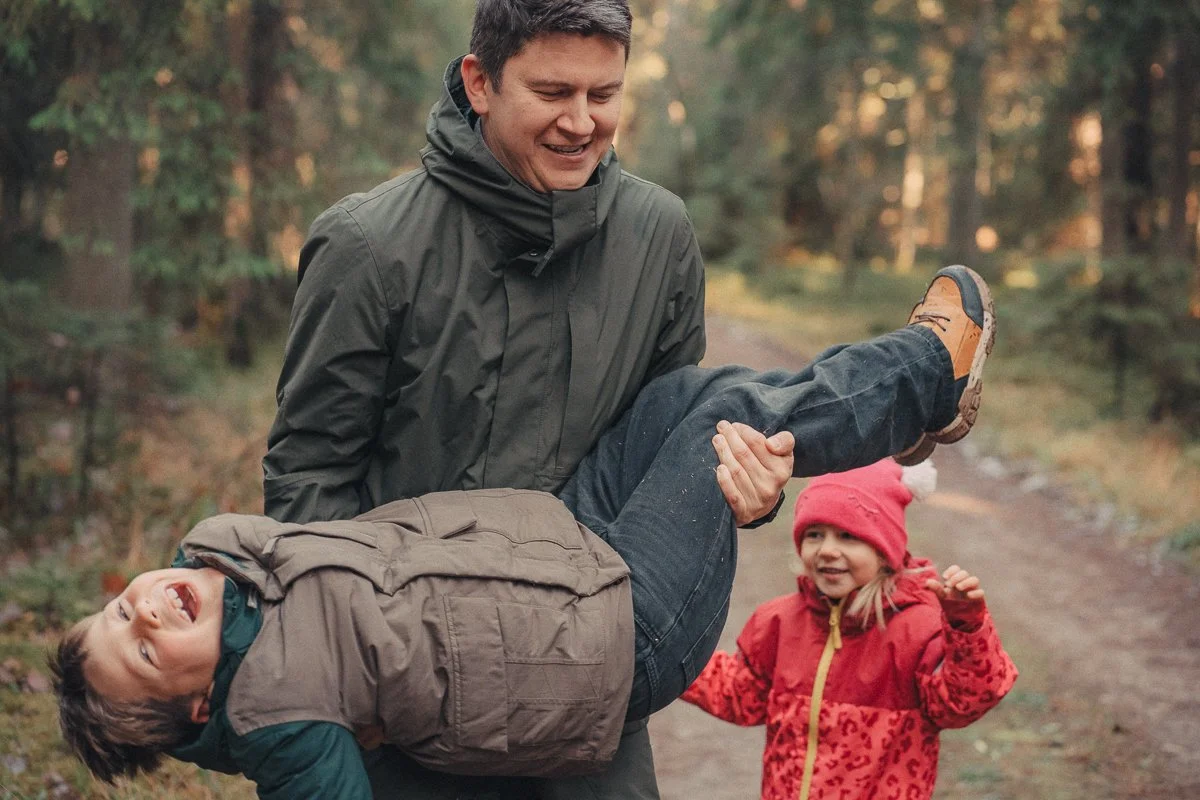 A man is smiling while carrying a young boy in his arms on a forest trail, and a girl in a red jacket and pink hat is standing nearby smiling.
