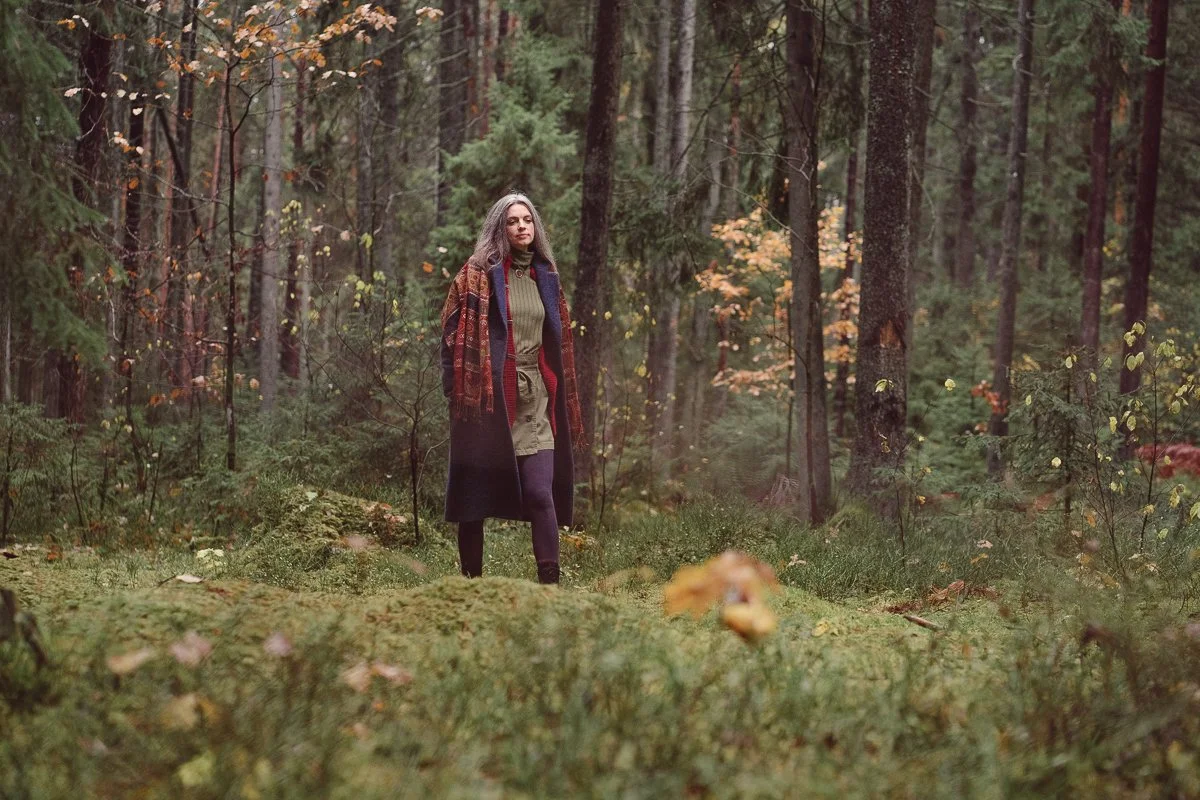 A woman walking through a dense forest with tall trees and green foliage, wearing a patterned coat over a green dress and black tights.