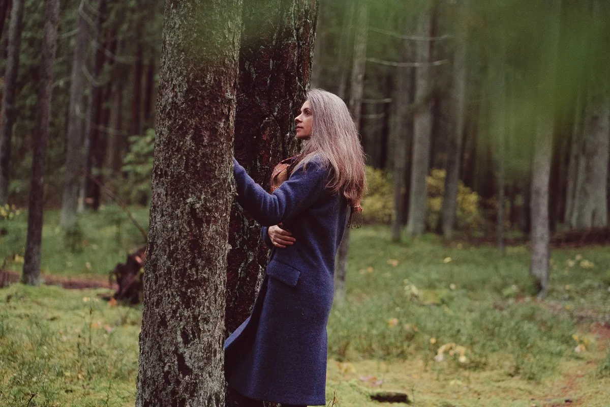 A woman with long gray hair stands hugging a tree in a forest, wearing a dark coat and gloves.