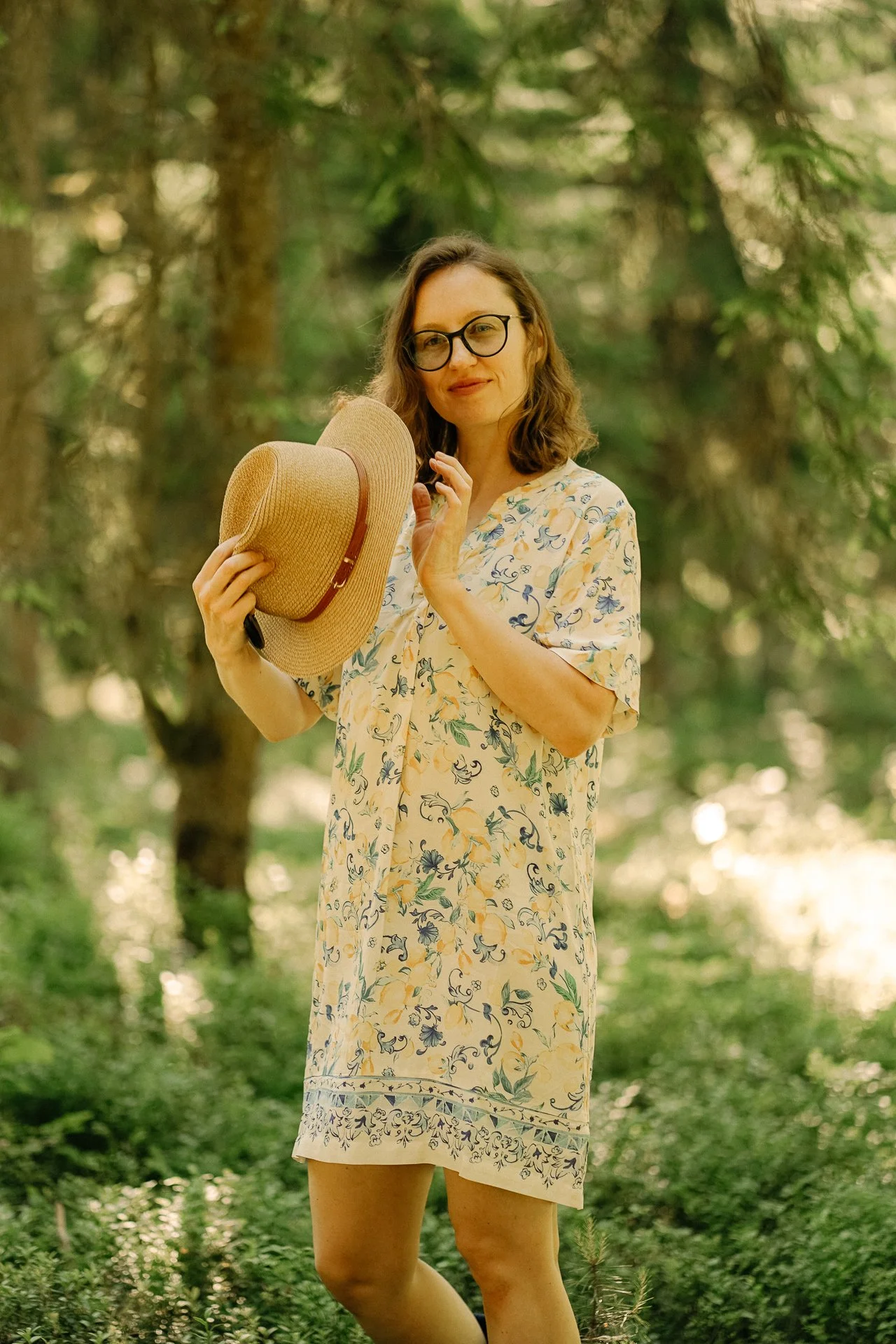 A woman in a floral dress holding a straw hat and standing outdoors in a lush green wooded area.