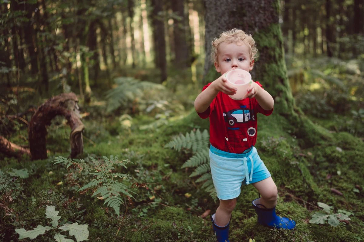 A young child with curly blonde hair in a red shirt and blue shorts is walking in a forest, holding a container with pink content, wearing blue rain boots.