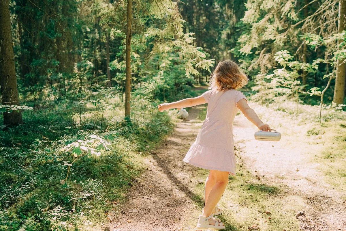 A young girl in a light pink dress twirling on a forest trail, holding a small white purse, surrounded by green trees and sunlight.