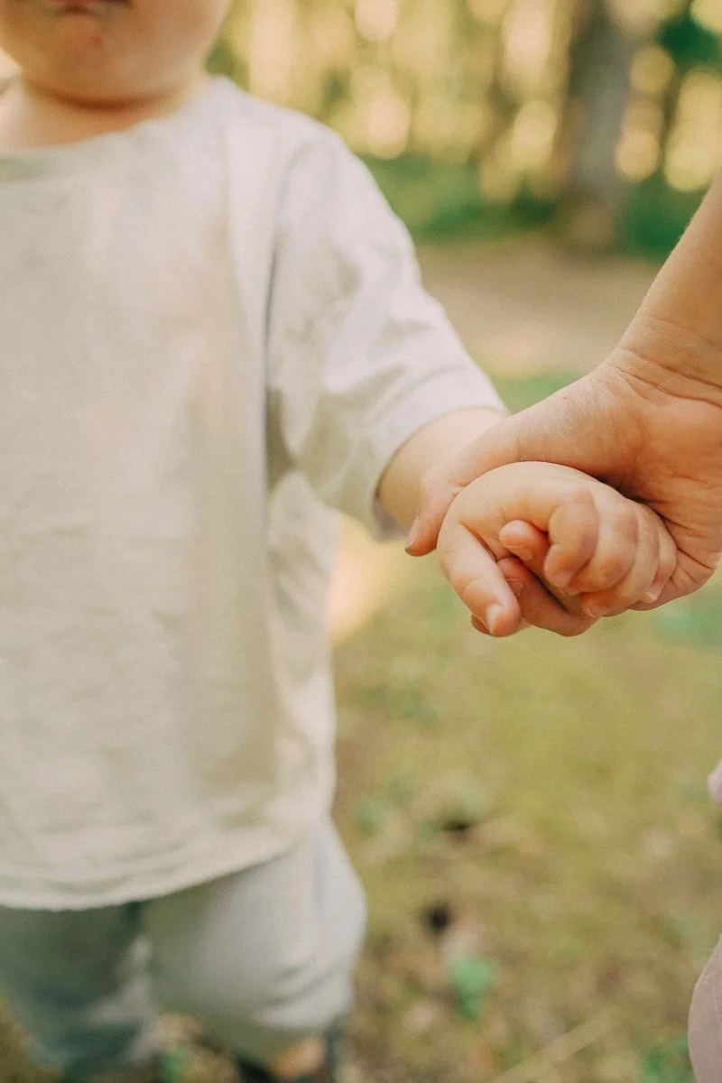 A young child holding an adult's hand outdoors with trees in the background.